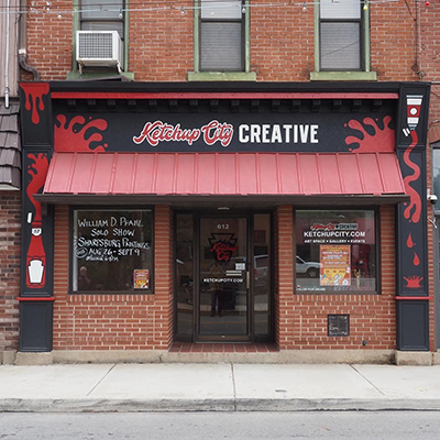 The exterior of a red brick building with artwork of ketchup bottles painted around the marquee and "Ketchup City Creative" written above the front door