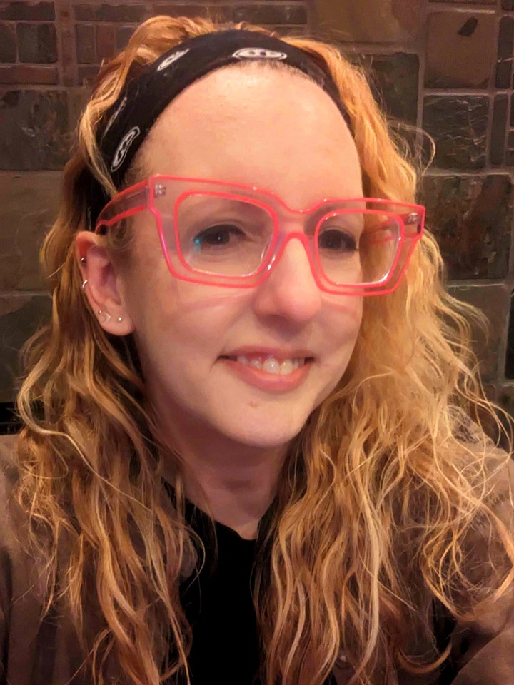 White woman with long blonde hair pulled back in a black bandana. She's wearing pink glasses, a black T-shirt, and a green shirt on top
