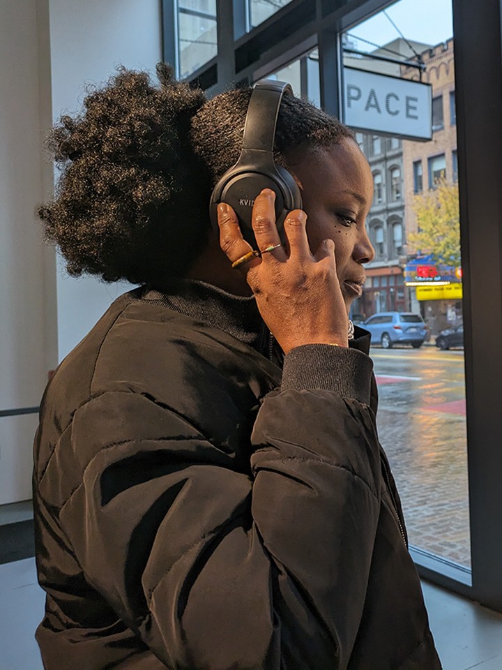 A Black woman wearing a black coat holds her hand up to her ear while listening to headphones inside an art gallery