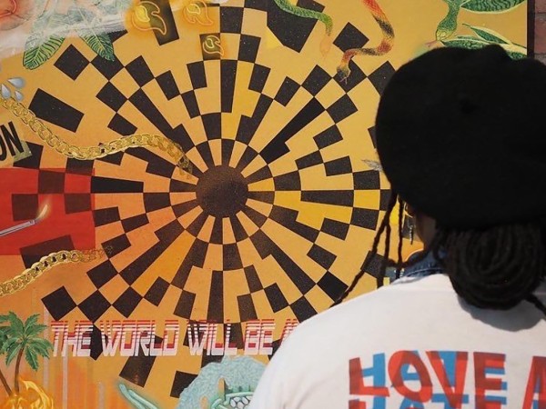 A person wearing a t-shirt that says "love me love me love me" on the back is shown from behind looking at a colorful piece of artwork hung on a brick wall