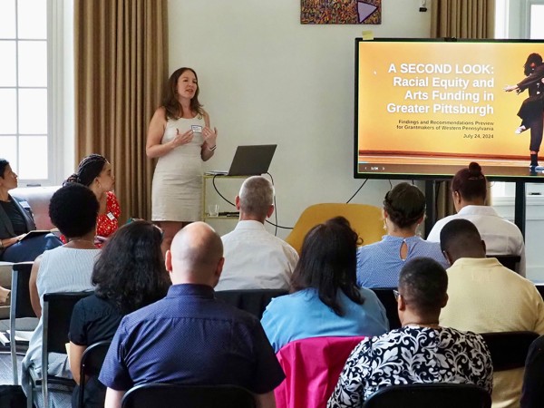 A crowd of people sit in folding chairs facing a white woman in front of a microphone addressing the crowd. To her left is a large monitor showing the cover of a report titled, "A Second Look: Racial Equity and Arts Funding in Greater Pittsburgh." To her right sits three women of the Global Majority sitting on a couch.