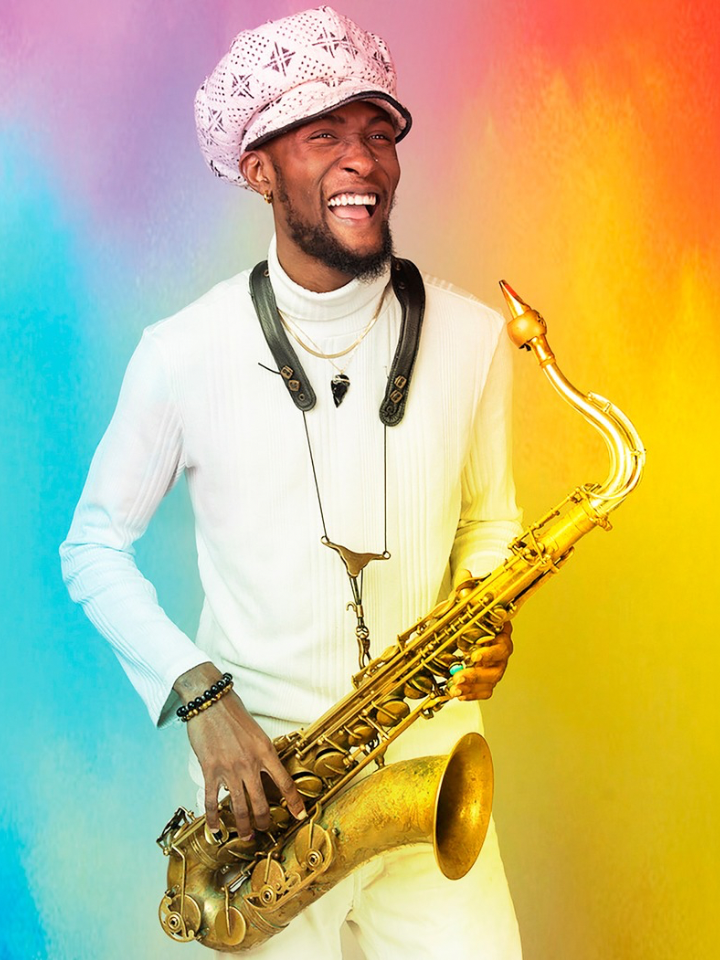 A smiling Black man with a close-cut dark beard wearing an all-white outfit, white hat, and a saxophone around his neck