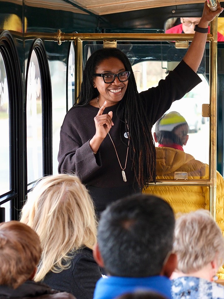 A smiling Black woman with black glasses and long dark hair stands in front of a trolley tour, speaking to a diverse group of seated people