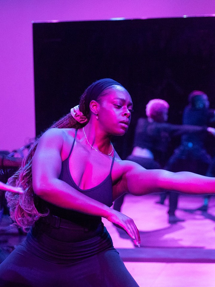 A purple-lit photo shows five Black dancers performing in front of a mirror