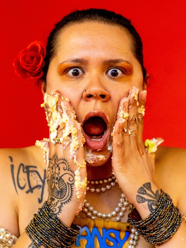 A black woman with a red flower in her hair making a shocked face. Her hands are up against her cheeks with cake on her fingers.