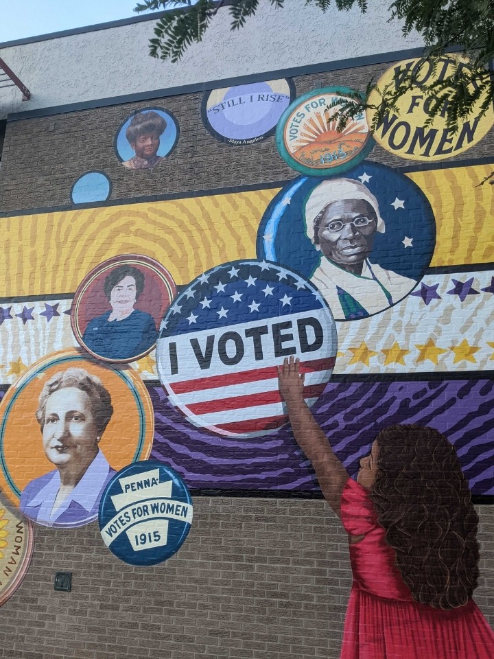 Close-up detail of a colorful mural showing a small dark-skinned child with black hair reaching up and touching an "I Voted" button, surrounded by other buttons featuring faces of diverse women