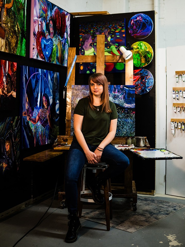 Portrait of a white woman with long straight dark blonde hair, wearing a black t-shirt and dark blue jeans, sitting on a stool surrounded by artwork