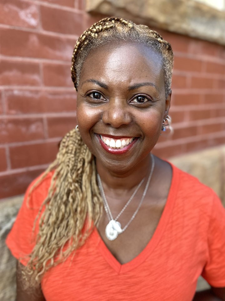 A smiling Black woman with long light colored hair poses for the camera. She's wearing neckalces and a v-neck orange shirt