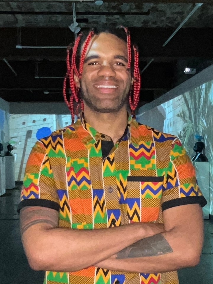 Smiling Black man with red-and-black shoulder-length braids and wearing a colorful African-inspired patterned shirt, poses for the camera while crossing his arms in front of an art gallery exhibit