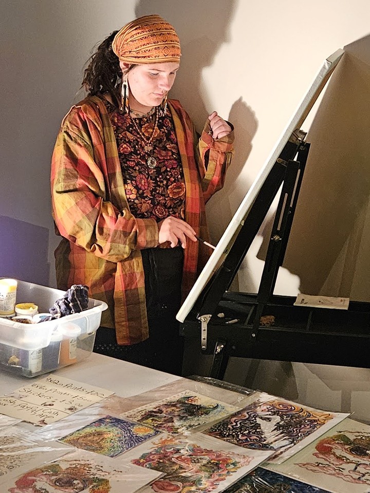 A person wearing bohemian-looking clothing and head scarf paints on an easel