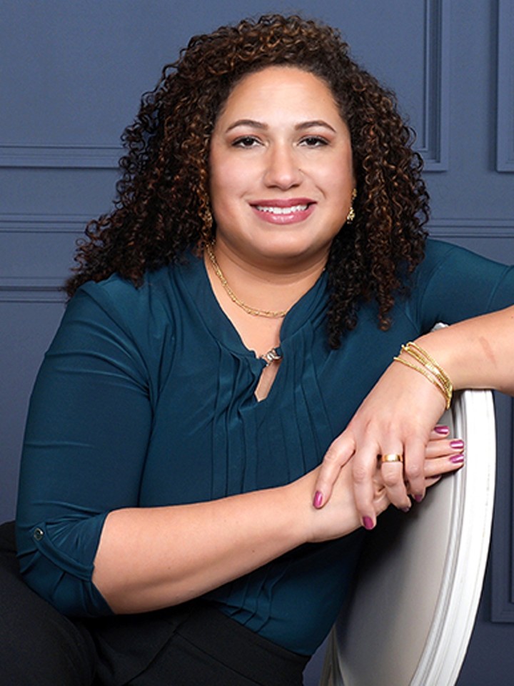 Portrait of Latine woman with curly dark hair, a blue long-sleeved shirt rolled up to her elbows, gold jewelry, and a big smile