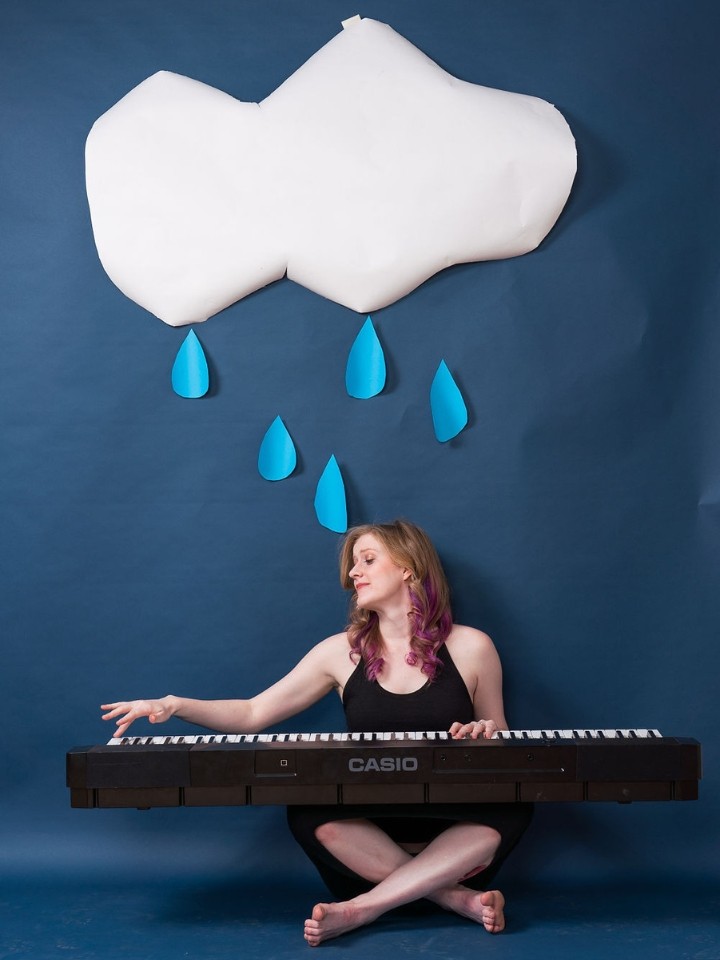 A white woman sits cross legged on a floor playing a large Casio keyboard below a paper-cut out of a large white cloud with blue raindrops