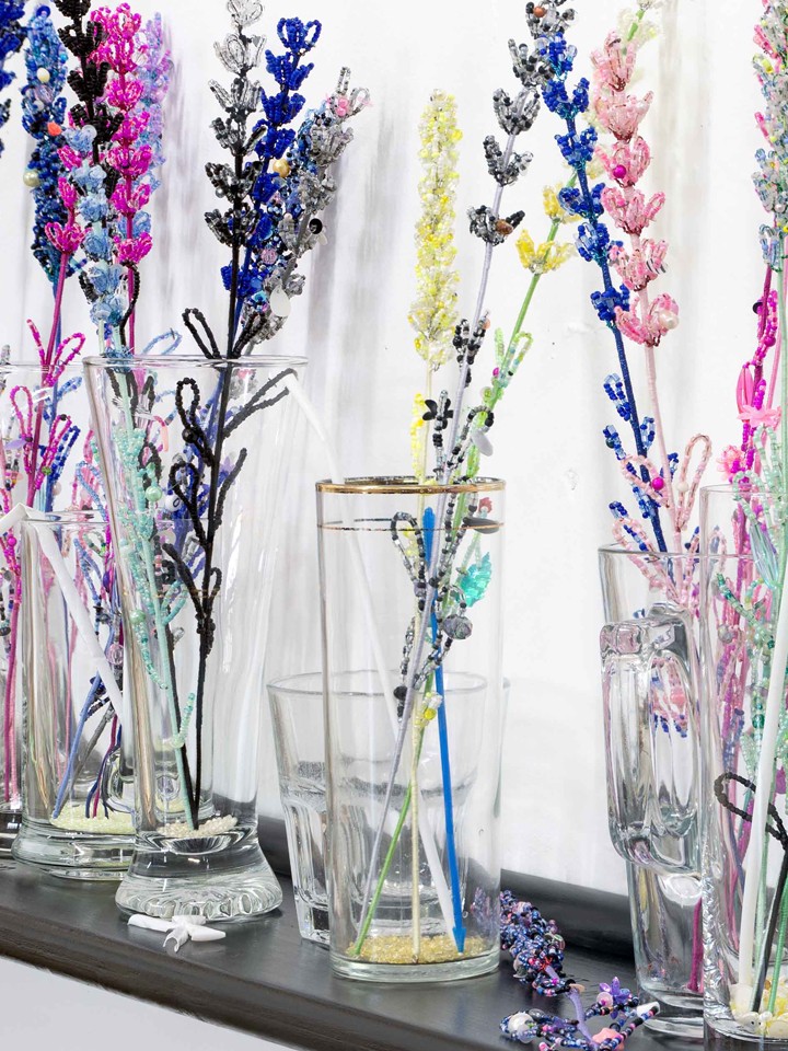 Colorful flower stems, created out of colorful beads, sit in glass jars on a black shelf inside a gallery space