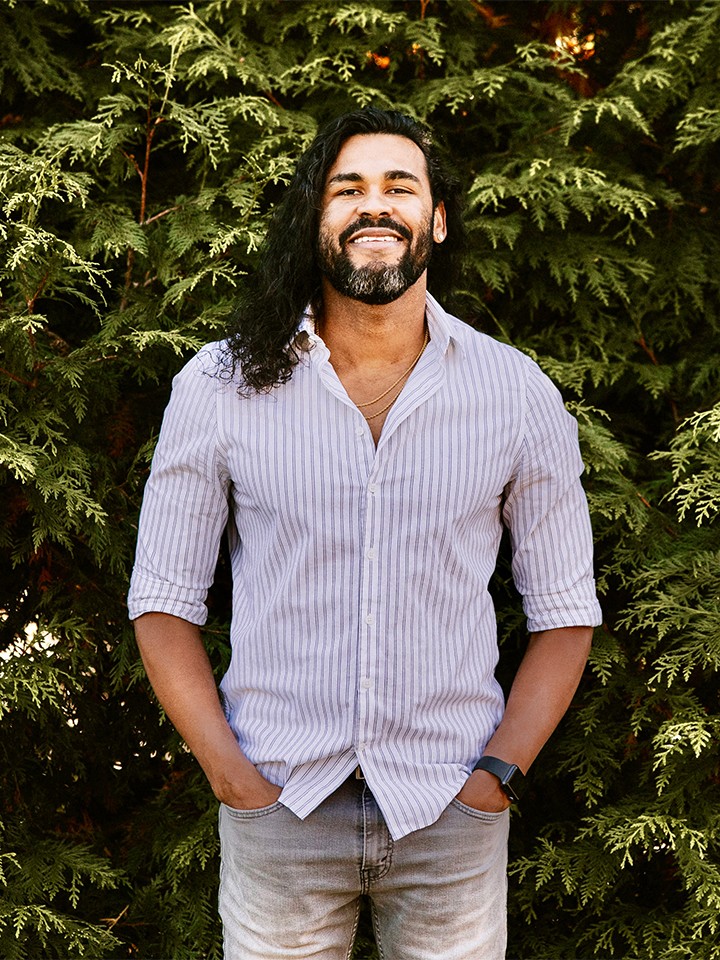 A smiling man with light brown skin, dark beard, and long dark hair, wearing a striped button down shirt, poses in front of green shrubbery