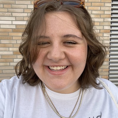 Smiling white woman with shoulder-length straight brown hair pulled back with brown sunglasses, wearing a white Fleetwood Mac shirt and several necklaces