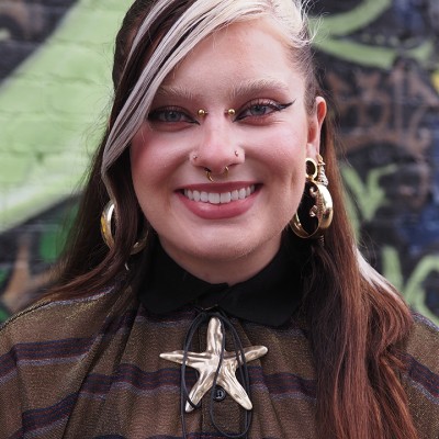 Smiling white person with long dark hair with chunky white streaks, multiple nose and ear piercings, wearing a brown striped shirt and a large metallic star necklace