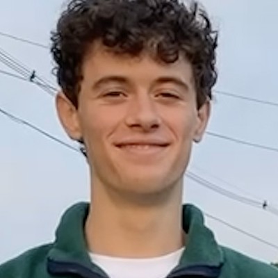 Portrait of a smiling white man with short dark curly hair, a white t-shirt, and a green zippered pullover