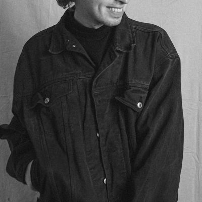 A black and white photo of Ryan Shimko, a young man with curly hair, wearing a dark jean jacket and glasses. Ryan is smiling while looking to the side.