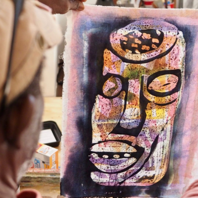 A person holds up a piece of artwork depicting a stylized abstract face in vibrant colors. The person holding the art is seen from behind, wearing a light brown cap, and is in an art studio, with supplies visible in the background.