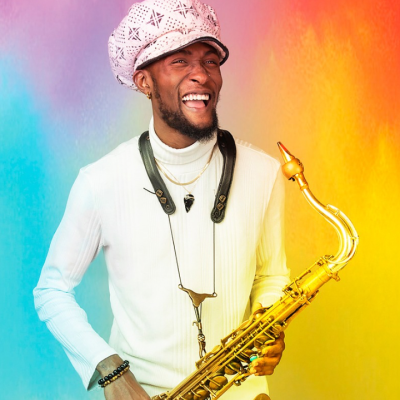 A smiling Black man with a close-cut dark beard wearing an all-white outfit, white hat, and a saxophone around his neck