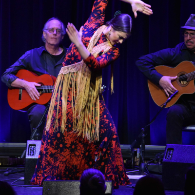 A person wearing a colorful dress and gold scarf dances in front of two people playing acoustic guitars