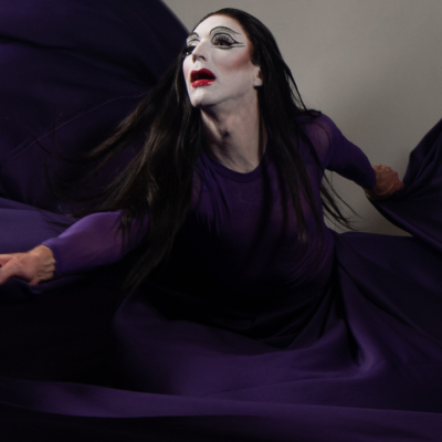 A person with long dark hair, white dramatic face makeup including black eyeliner and red lipstick, poses while holding onto a long purple gown