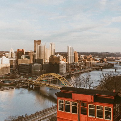 A photo of Pittsburgh's skyline, a yellow bridge, and a river, with a red trolley visible in the foreground