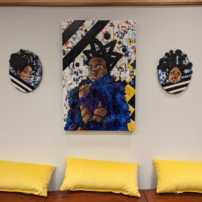 Three pieces of artwork showing Black women hang on a wall above a wooden bench and yellow pillows