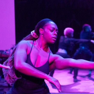 A purple-lit photo shows five Black dancers performing in front of a mirror