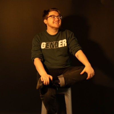 A white person with short hair, glasses sitting on a stool with a shirt that says gender 