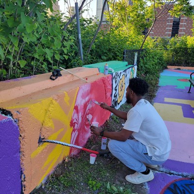 A Black person paints a colorful mural in an outside graffiti park. A view of Downtown Pittsburgh is visible in the background