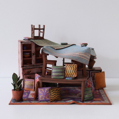 A sculpture depicting a miniature messy dining room scene, including boxes and stools sitting on top of a wooden table