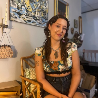 A white woman with long brown hair wearing a floral shirt and a pair of short dark shorts smile for the camera while sitting in a chair