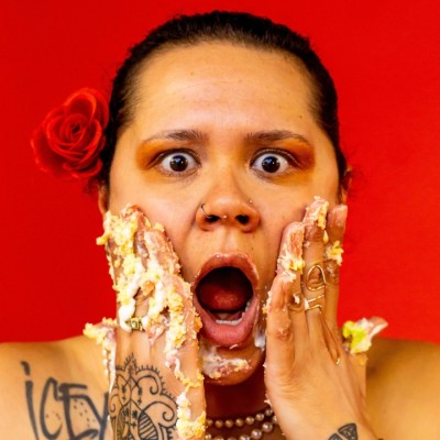 A black woman with a red flower in her hair making a shocked face. Her hands are up against her cheeks with cake on her fingers.