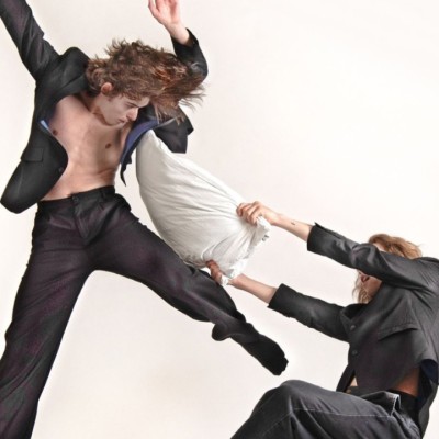 Two white male dancers wearing black suits as one leaps away from the other who is hitting him with a pillow