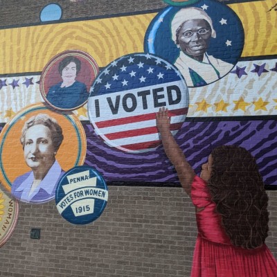 Close-up detail of a colorful mural showing a small dark-skinned child with black hair reaching up and touching an "I Voted" button, surrounded by other buttons featuring faces of diverse women