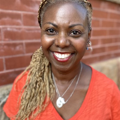 A smiling Black woman with long light colored hair poses for the camera. She's wearing neckalces and a v-neck orange shirt