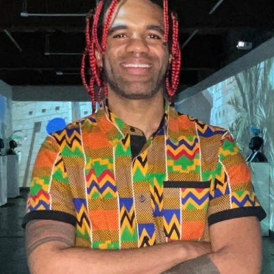 Smiling Black man with red-and-black shoulder-length braids and wearing a colorful African-inspired patterned shirt, poses for the camera while crossing his arms in front of an art gallery exhibit