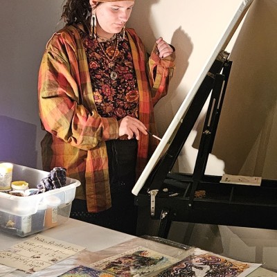 A person wearing bohemian-looking clothing and head scarf paints on an easel