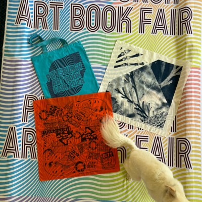 a colorful banner that reads "Pittsburgh Art Book Fair" in bold text. The background of the banner has a wavy, rainbow-striped pattern. On top of the banner, there are several items displayed, including a teal tote bag with "Pittsburgh Art Book Fair 2024" printed on it, an orange cloth or poster with various graphic designs, and a light-colored fabric with botanical prints. A small, white dog is standing on the banner, partially covering some of the text.