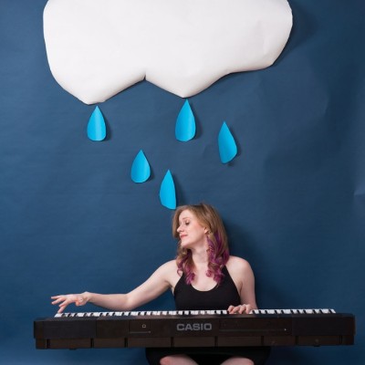 A white woman sits cross legged on a floor playing a large Casio keyboard below a paper-cut out of a large white cloud with blue raindrops