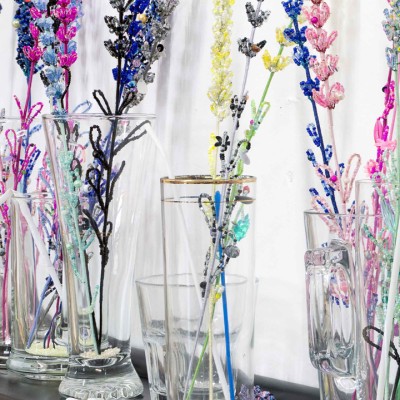 Colorful flower stems, created out of colorful beads, sit in glass jars on a black shelf inside a gallery space