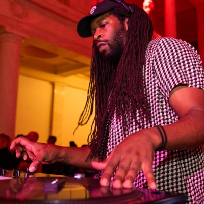 A Black man with a dark beard, long dark hair, and a baseball cap is shown spinning records on a turntable inside an art space