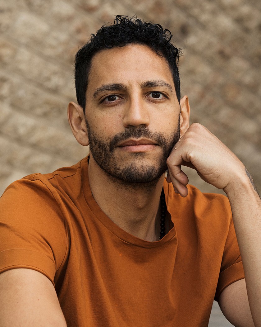 A man with light brown skin, dark beard, and short dark hair holds up his hand to his cheek in pose