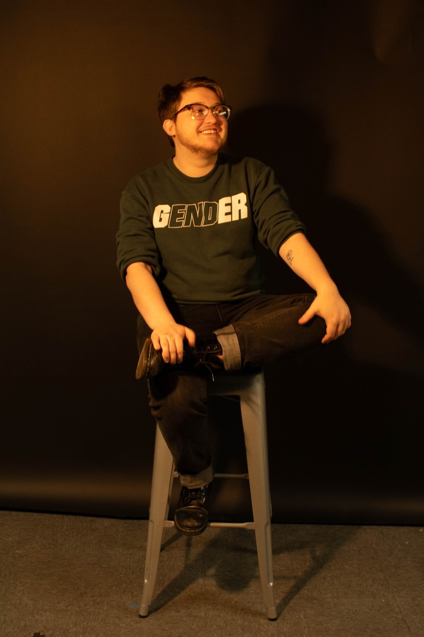 A white person with short hair, glasses sitting on a stool with a shirt that says gender 