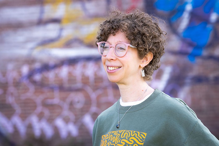 A person with light skin and short curly dark hair, pink glasses, and a green sweatshirt featuring a brightly colored yellow abstract design on its front smiles in front of a graffiti wall