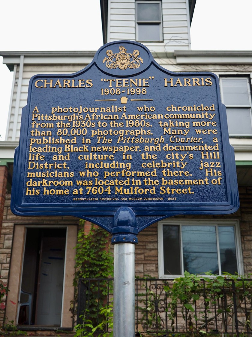 A blue sign with yellow text is mounted on a silver sign pole in front of a row house. The sign says Charles "Teenie" Harris