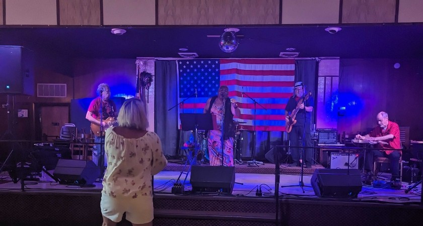 A stage with and American flag and blue lights and four people playing instruments, with a blonde haired woman facing them 