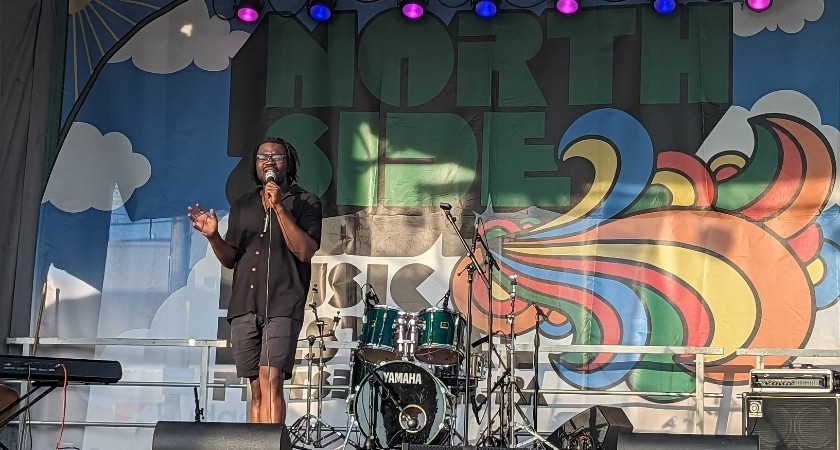 A black man on a stage with a drumset behind him and a banner that says North Side