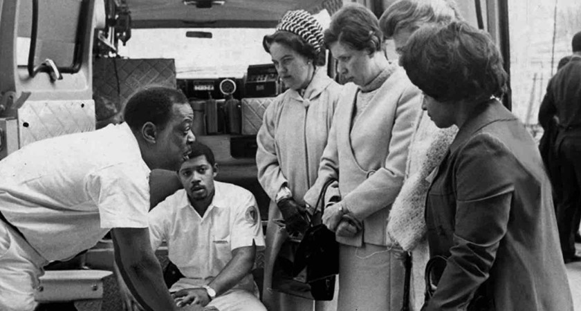 A black and white photo of two black male paramedics speaking to four women